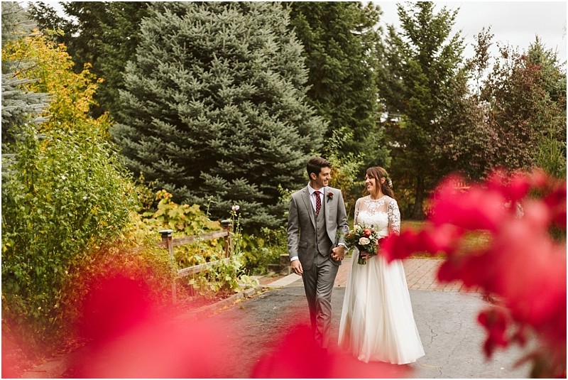 Premier Snohomish & Seattle Wedding Photographer | GSquared Weddings Photography A bride and groom walk hand in hand down a garden path, smiling at each other, surrounded by lush greenery and vibrant red flowers—capturing the romance of a Snohomish Seattle wedding engagement. 47.9129° N, 122.0982° W | Serving Snohomish, Seattle , Orlando & Beyond