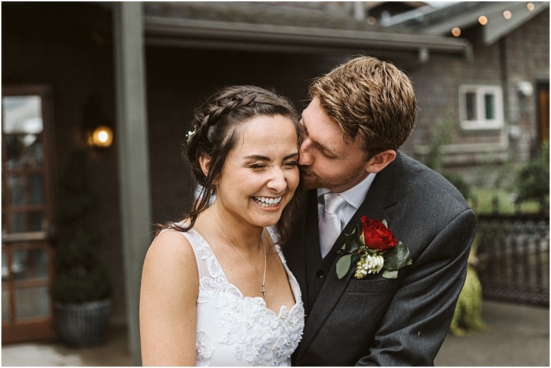 Premier Snohomish & Seattle Wedding Photographer | GSquared Weddings Photography A groom in a suit kisses a smiling bride on the cheek during their joyful Snohomish outdoor wedding, both appearing affectionate. The bride wears a white dress and the groom highlights his look with a red rose boutonniere. 47.9129° N, 122.0982° W | Serving Snohomish, Seattle , Orlando & Beyond