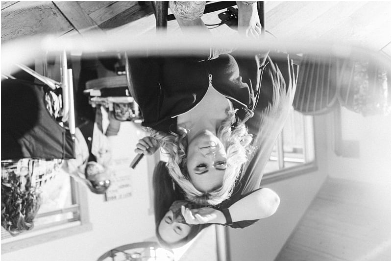 Black and white photo of a woman in Snohomish getting her hair styled with a curling iron, reflected upside down in a mirror. Other people and furniture are visible in the background. Snohomish Wedding Photography