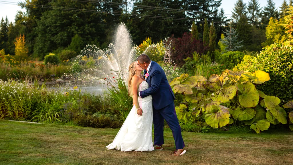 A bride and groom share a kiss outdoors on a lush green lawn at one of the picturesque Snohomish wedding venues with a fountain and vibrant garden plants in the background on a sunny day Snohomish Wedding Photography A bride and groom share a kiss outdoors on a lush green lawn at one of the picturesque Snohomish wedding venues with a fountain and vibrant garden plants in the background on a sunny day Snohomish Wedding Photography