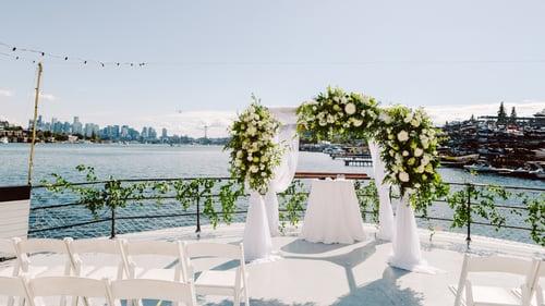 A wedding arch decorated with white flowers and greenery stands on a boat deck overlooking a city skylineone of the most unique Washington wedding venueswhite chairs await guests with sparkling water in the background under a sunny sky Snohomish Wedding Photography A wedding arch decorated with white flowers and greenery stands on a boat deck overlooking a city skylineone of the most unique Washington wedding venueswhite chairs await guests with sparkling water in the background under a sunny sky Snohomish Wedding Photography