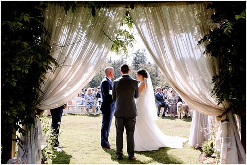 A bride and groom stand facing each other at an outdoor ceremony framed by sheer curtains and greenery with guests on white chairs Many Snohomish wedding venues offer similar picturesque settings for your special day Snohomish Wedding Photography A bride and groom stand facing each other at an outdoor ceremony framed by sheer curtains and greenery with guests on white chairs Many Snohomish wedding venues offer similar picturesque settings for your special day Snohomish Wedding Photography