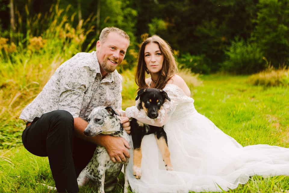GSquared Weddings Photography A couple poses outdoors on grass, the woman in a white dress and the man in a patterned shirt. They are each holding a dog, surrounded by greenery and tall grass. 47.9129° N, 122.0982° W | Serving Snohomish, Seattle , Orlando & Beyond