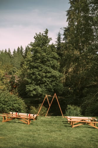 A grassy outdoor area with wooden benches arranged on both sides facing a simple triangular wooden arch surrounded by lush green trees under a partly cloudy sky Snohomish Wedding Photography A grassy outdoor area with wooden benches arranged on both sides facing a simple triangular wooden arch surrounded by lush green trees under a partly cloudy sky Snohomish Wedding Photography