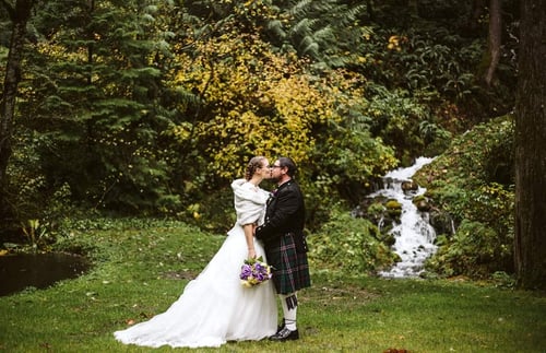 A bride in a white gown and groom in a kilt embrace outdoors in front of trees and a small waterfall with the bride holding a bouquet of purple flowers Snohomish Wedding Photography A bride in a white gown and groom in a kilt embrace outdoors in front of trees and a small waterfall with the bride holding a bouquet of purple flowers Snohomish Wedding Photography