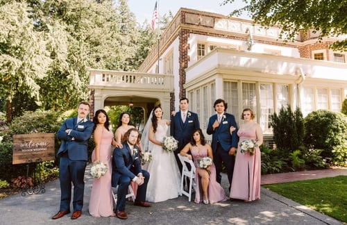 A wedding party poses outdoors in front of a large elegant house The bride stands in the center holding a bouquet surrounded by bridesmaids in pink dresses and groomsmen in blue suits Greenery and a welcome sign are visible Snohomish Wedding Photography A wedding party poses outdoors in front of a large elegant house The bride stands in the center holding a bouquet surrounded by bridesmaids in pink dresses and groomsmen in blue suits Greenery and a welcome sign are visible Snohomish Wedding Photography