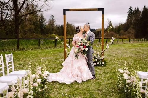 A couple embraces and kisses at an outdoor wedding ceremony standing in front of a wooden arch decorated with flowers The bride wears a pink floral gown and the groom is in a light gray suit White chairs and flowers line the aisle Snohomish Wedding Photography A couple embraces and kisses at an outdoor wedding ceremony standing in front of a wooden arch decorated with flowers The bride wears a pink floral gown and the groom is in a light gray suit White chairs and flowers line the aisle Snohomish Wedding Photography