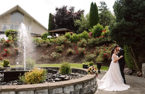 A bride and groom embrace and kiss beside a fountain in a landscaped garden with a bouquet on the ground and a large house in the background surrounded by greenery and colorful flowers Snohomish Wedding Photography A bride and groom embrace and kiss beside a fountain in a landscaped garden with a bouquet on the ground and a large house in the background surrounded by greenery and colorful flowers Snohomish Wedding Photography