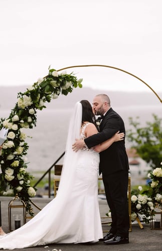 A bride and groom share a kiss under a circular floral arch at their outdoor wedding ceremony with water and greenery visible in the background Snohomish Wedding Photography A bride and groom share a kiss under a circular floral arch at their outdoor wedding ceremony with water and greenery visible in the background Snohomish Wedding Photography