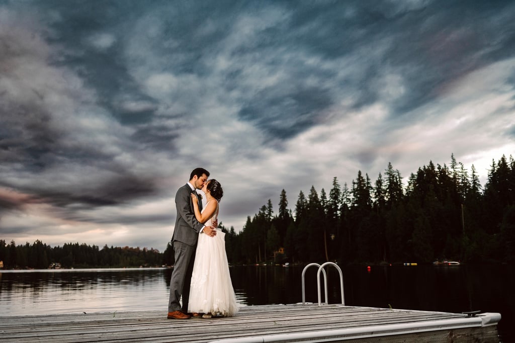 A bride and groom embrace on a wooden dock over a calm lake surrounded by tall trees and under a dramatic cloudy sky at dusk Snohomish Wedding Photography A bride and groom embrace on a wooden dock over a calm lake surrounded by tall trees and under a dramatic cloudy sky at dusk Snohomish Wedding Photography