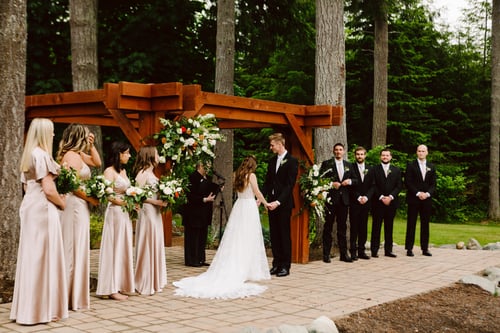 A bride and groom stand under a wooden arbor during an outdoor wedding ceremony holding hands Bridesmaids in champagne dresses and groomsmen in black suits stand on either side surrounded by tall trees and greenery Snohomish Wedding Photography A bride and groom stand under a wooden arbor during an outdoor wedding ceremony holding hands Bridesmaids in champagne dresses and groomsmen in black suits stand on either side surrounded by tall trees and greenery Snohomish Wedding Photography