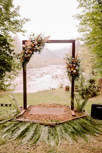 A wooden wedding arch decorated with pink and white flowers and greenery stands outdoors by a river surrounded by lush trees and ferns creating a natural and scenic ceremony setting Snohomish Wedding Photography A wooden wedding arch decorated with pink and white flowers and greenery stands outdoors by a river surrounded by lush trees and ferns creating a natural and scenic ceremony setting Snohomish Wedding Photography