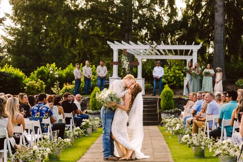 A bride and groom kiss at the outdoor altar during their wedding ceremony surrounded by guests seated on either side of a flowerlined aisle with the wedding party standing in the background Snohomish Wedding Photography A bride and groom kiss at the outdoor altar during their wedding ceremony surrounded by guests seated on either side of a flower lined aisle with the wedding party standing in the background Snohomish Wedding Photography
