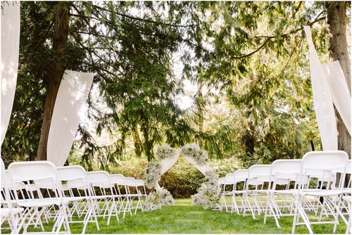 Hitching Post wedding venue Hitching Post wedding venue Rows of white folding chairs face a floral archway outdoors set on green grass beneath tall trees with flowing white drapes hanging between trunks creating a serene wedding ceremony setting Snohomish Wedding Photography