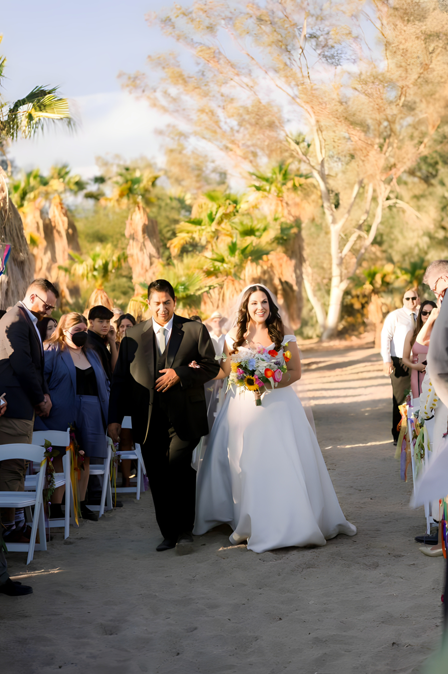 Bride being walked down the sandy aisle with colorful bouquet at Bubbling Wells Oasis