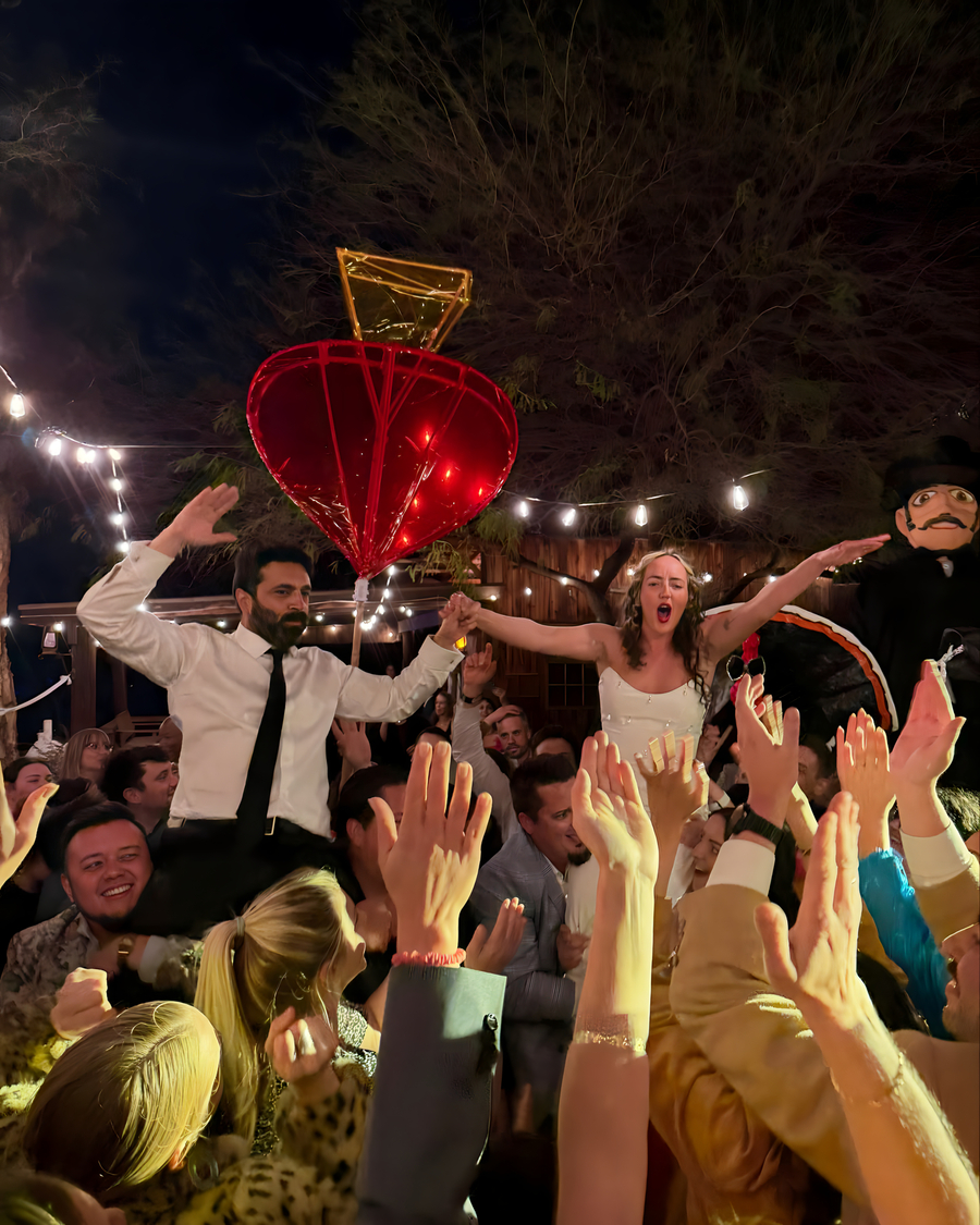 Bride and groom lifted by crowd of cheering guests at outdoor wedding reception