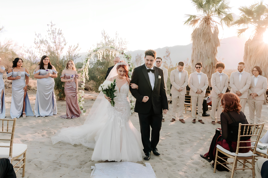 Couple walking up aisle with floral arch and bridesmaids in lavender dresses