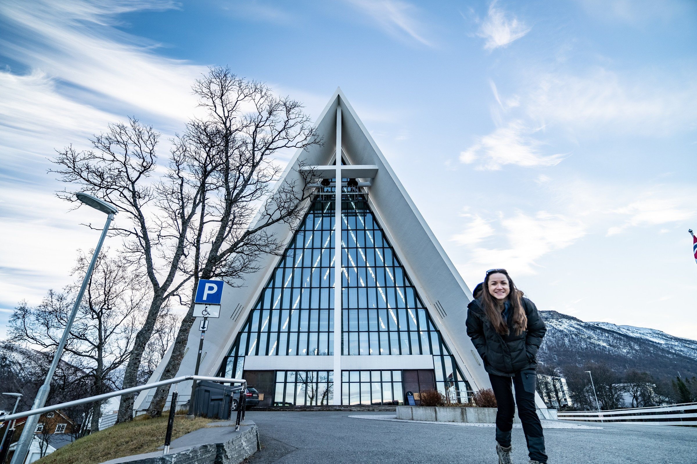 Wisatawan berjaket berdiri di depan Gereja Arktik Tromso dengan pegunungan bersalju di belakang