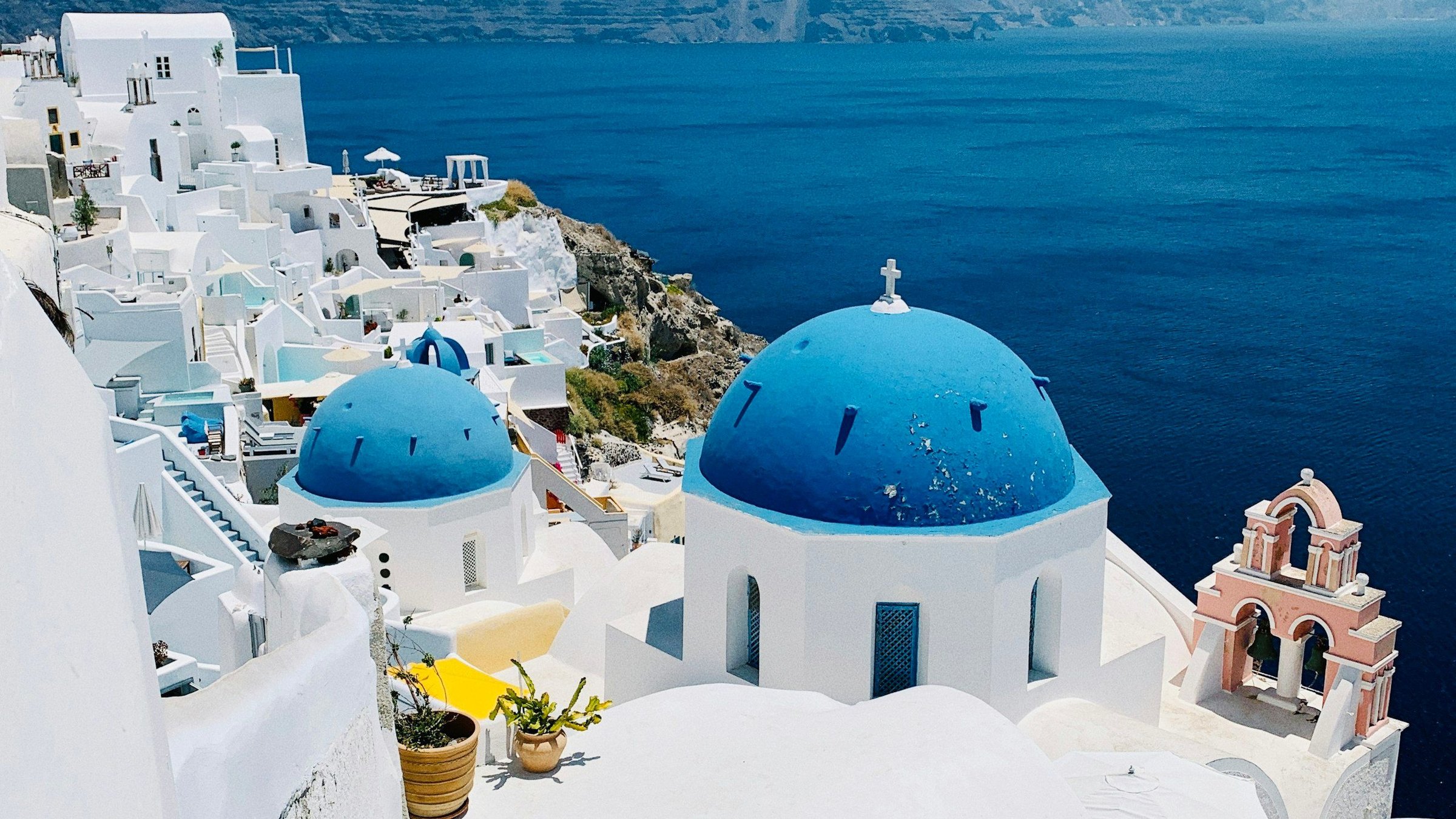 Kubah biru ikonik dan rumah putih desa Oia di Santorini menghadap laut Aegean dengan menara lonceng merah muda di bawah langit musim panas yang cerah