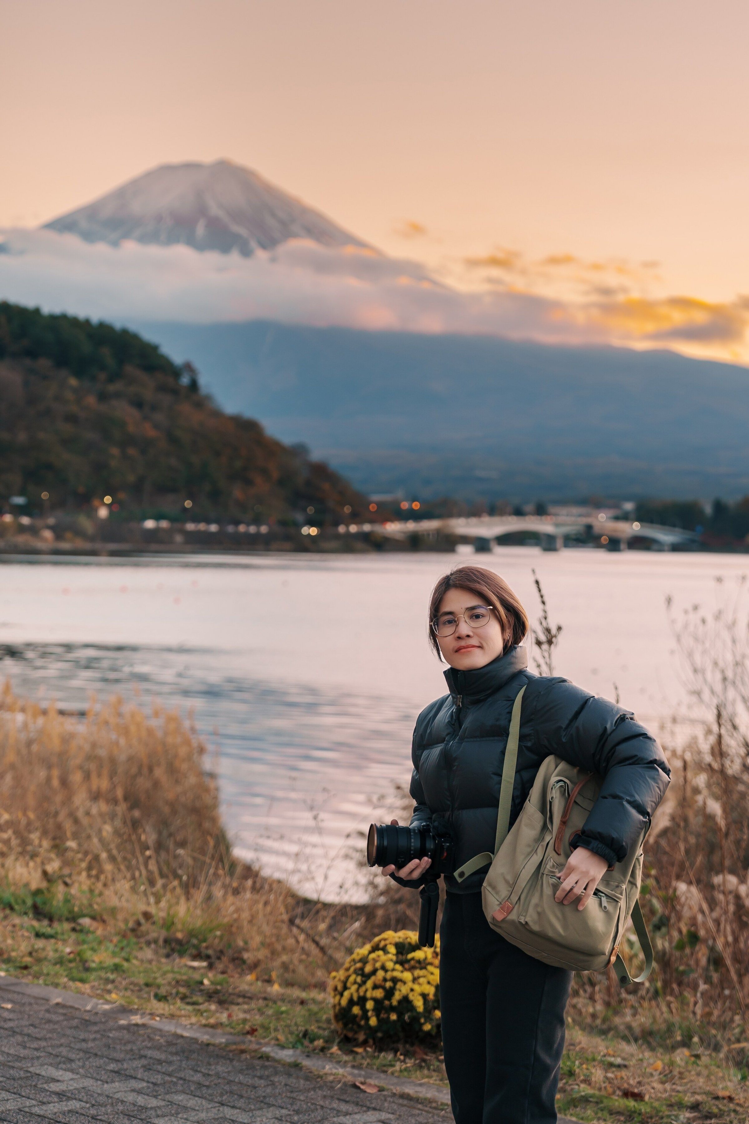 Wisatawan berjaket puffer membawa kamera di tepi Danau Kawaguchi dengan Gunung Fuji dan langit senja jingga