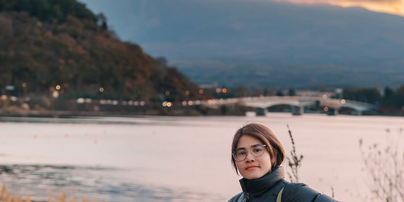 Wisatawan berjaket puffer membawa kamera di tepi Danau Kawaguchi dengan Gunung Fuji dan langit senja jingga