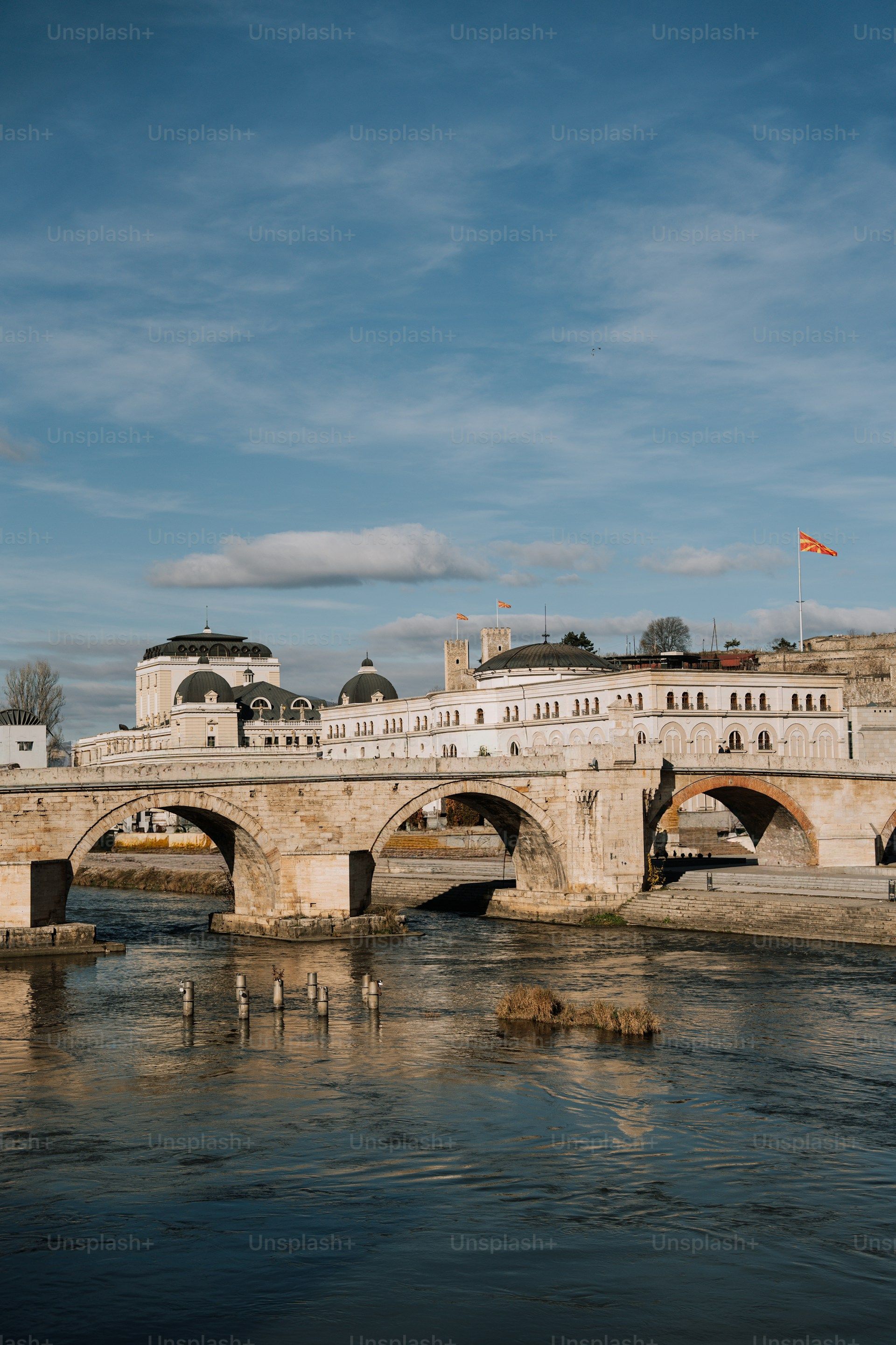 Jembatan Batu Kameni Most di atas Sungai Vardar dengan gedung plaza putih dan bendera Makedonia Utara, Skopje