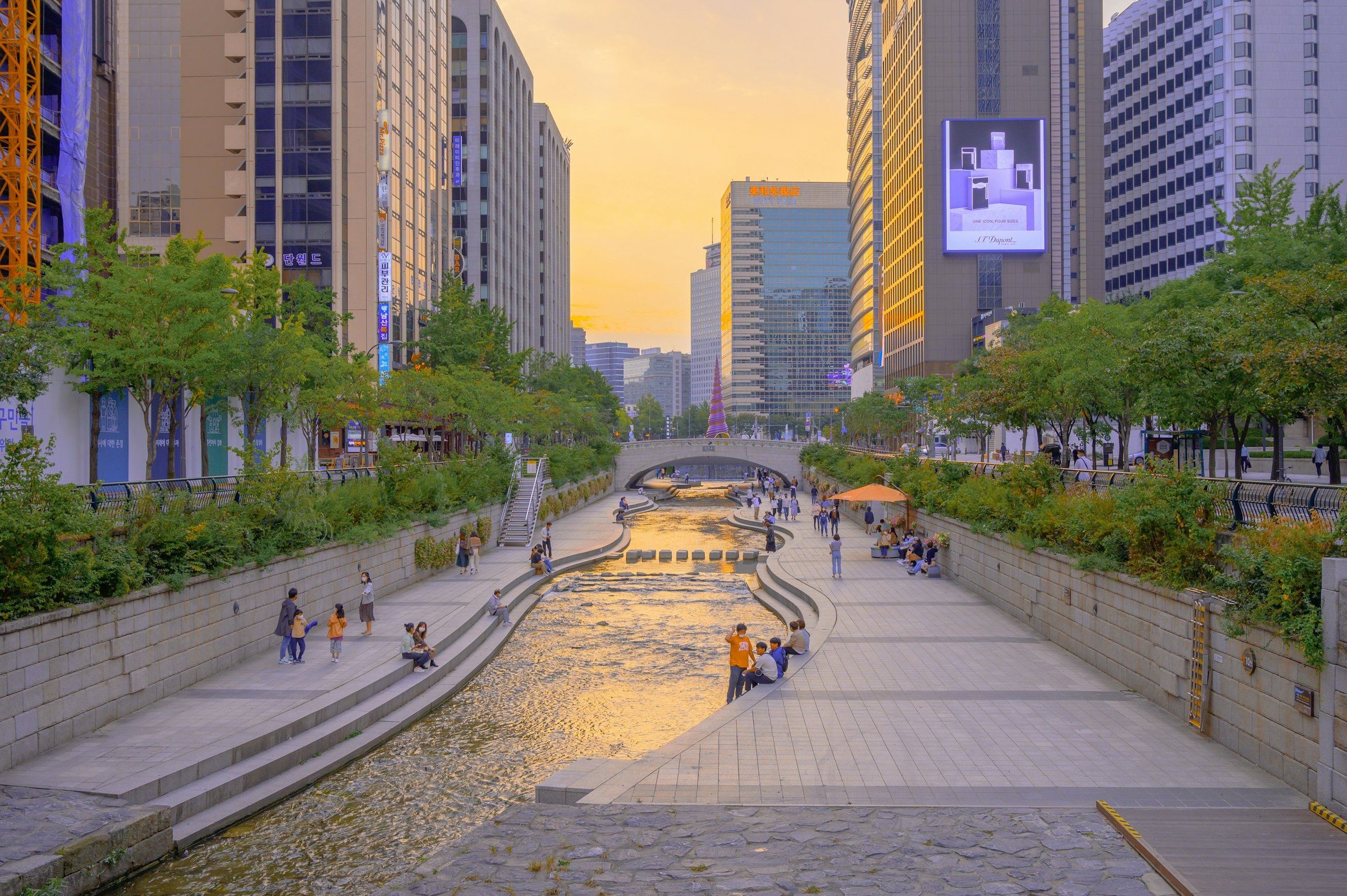 Taman Cheonggyecheon di Seoul saat senja, orang berjalan di tepi sungai dengan gedung pencakar langit