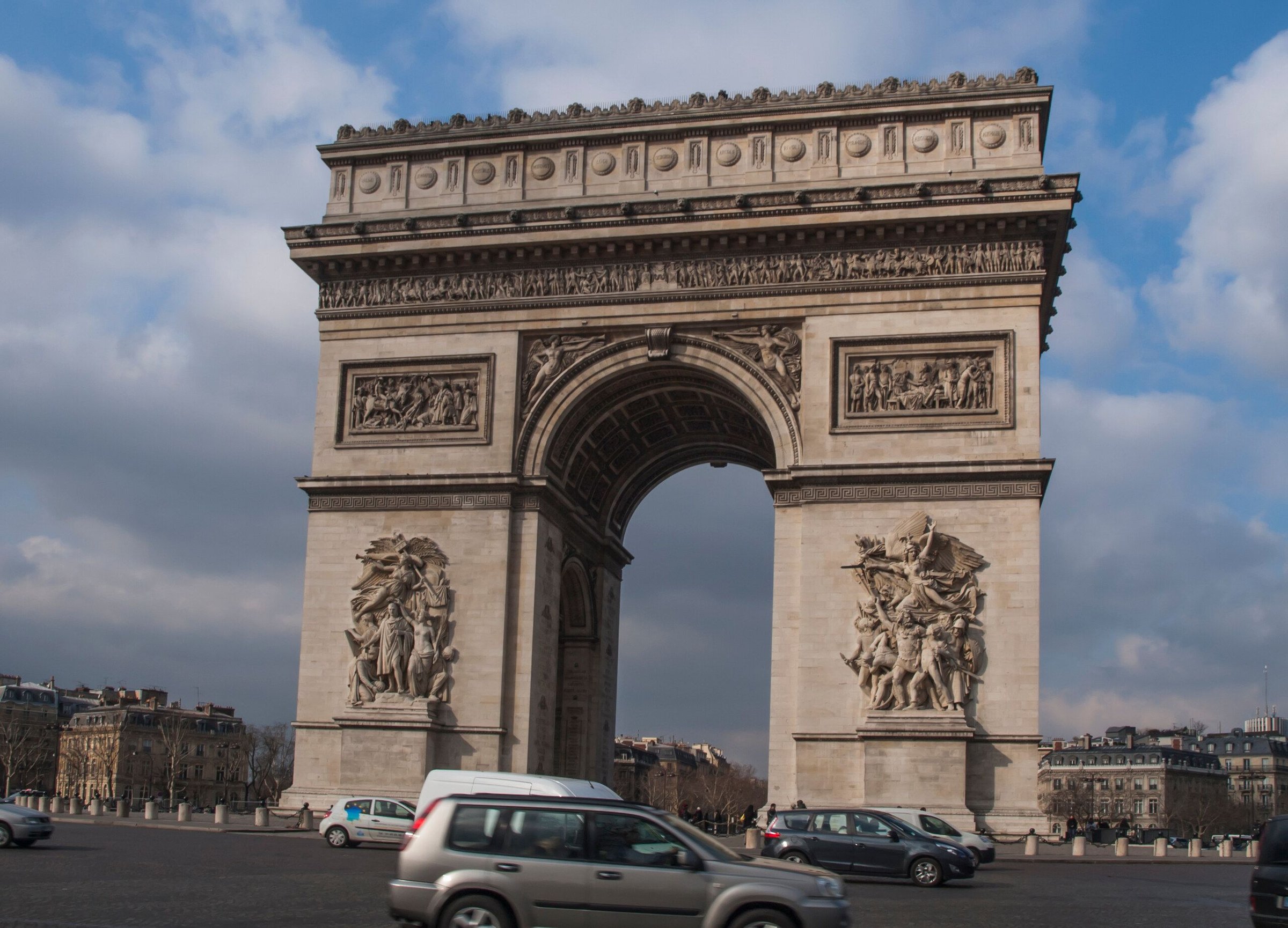 Arc de Triomphe di Paris dengan langit berawan dan mobil melintas di bawahnya.