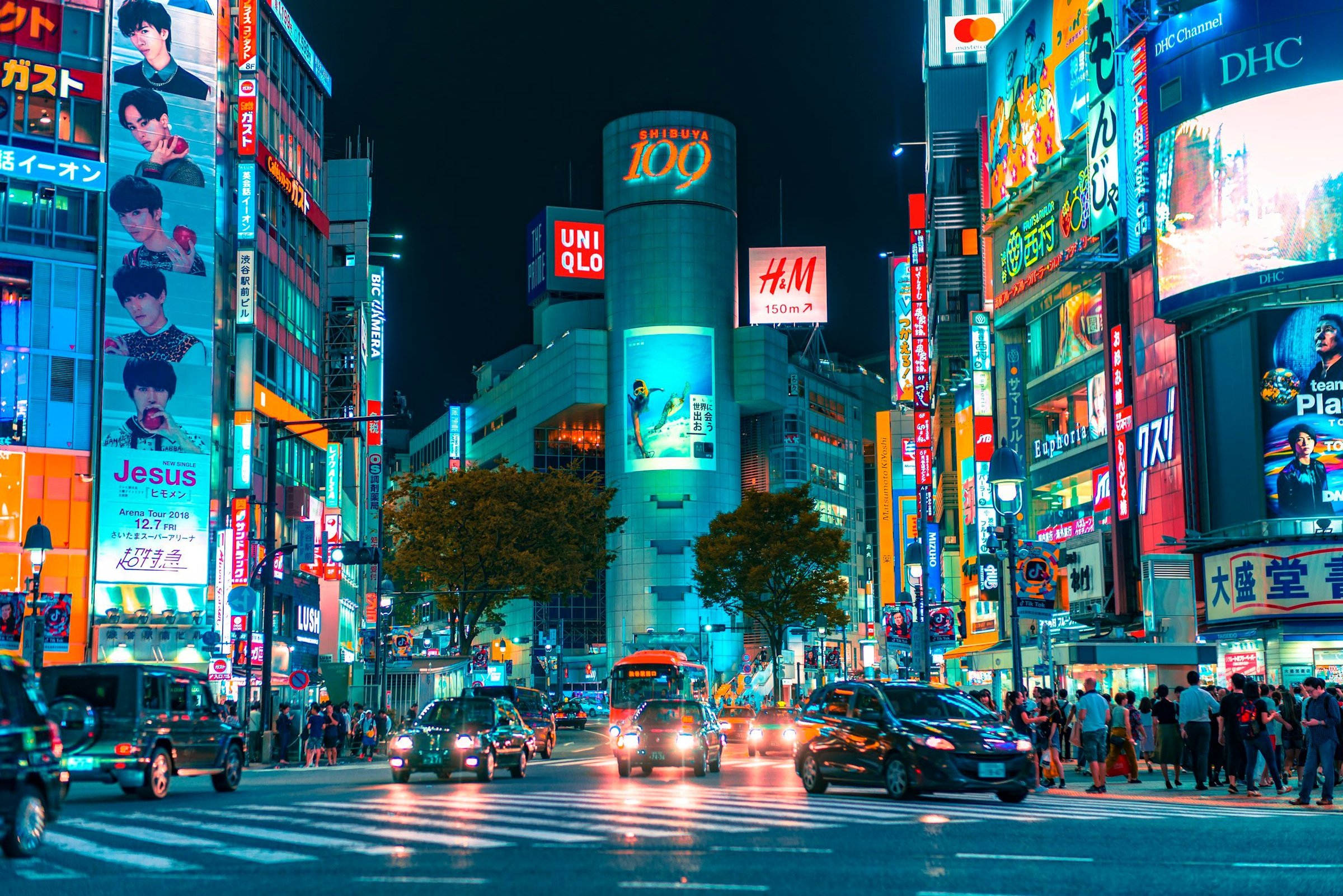 Persimpangan Shibuya, Tokyo, pada malam hari dengan gedung SHIBUYA 109, papan reklame neon, dan mobil melintasi pedestrian crossing yang sibuk