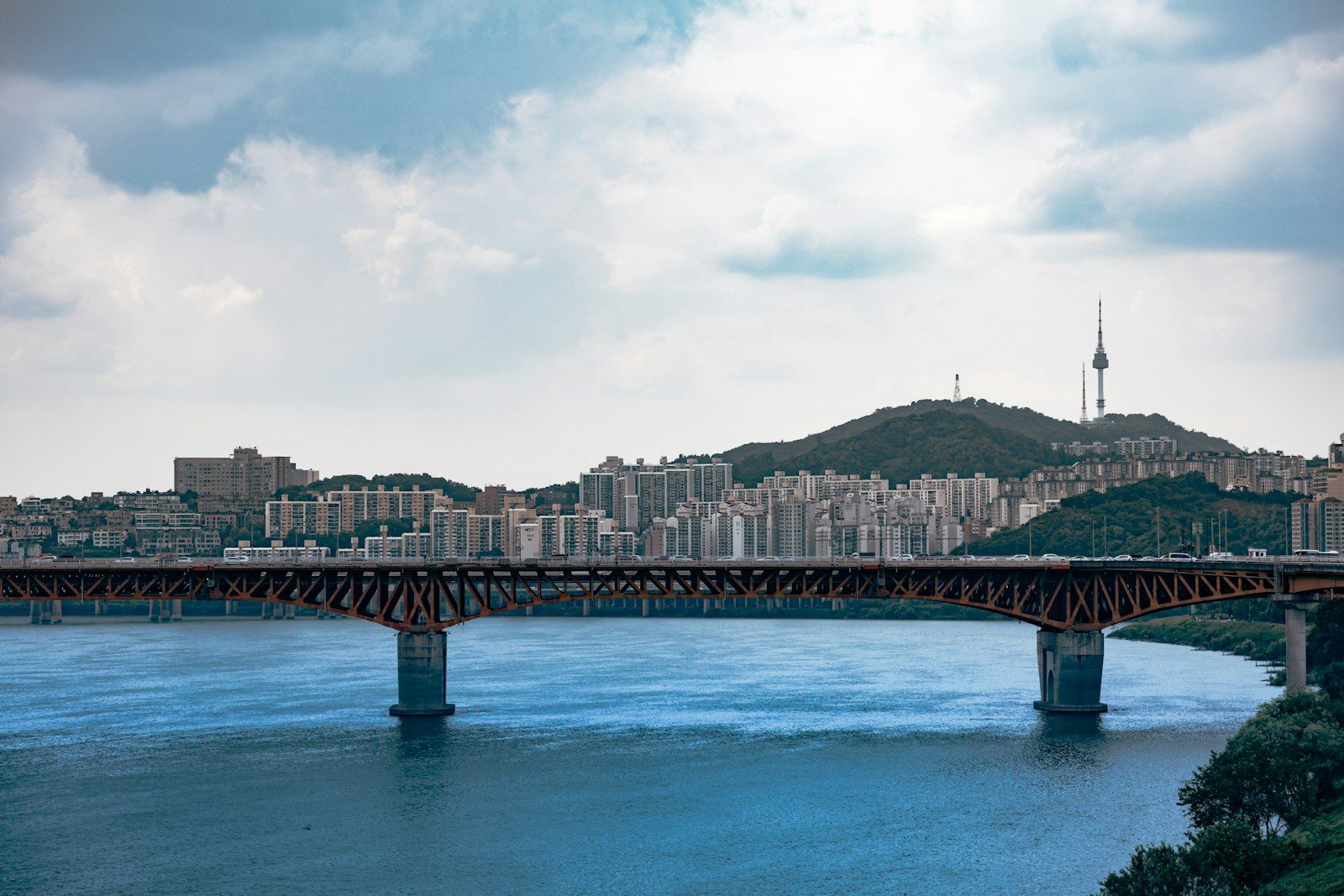 Jembatan oranye membentang di atas sungai, pemandangan kota dan Menara Namsan di perbukitan.