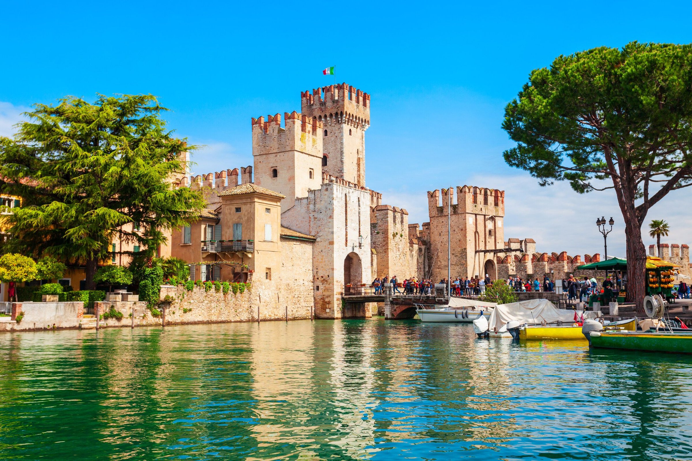 Kastil Scaligero Sirmione dilihat dari seberang air danau dengan pohon pinus dan perahu.