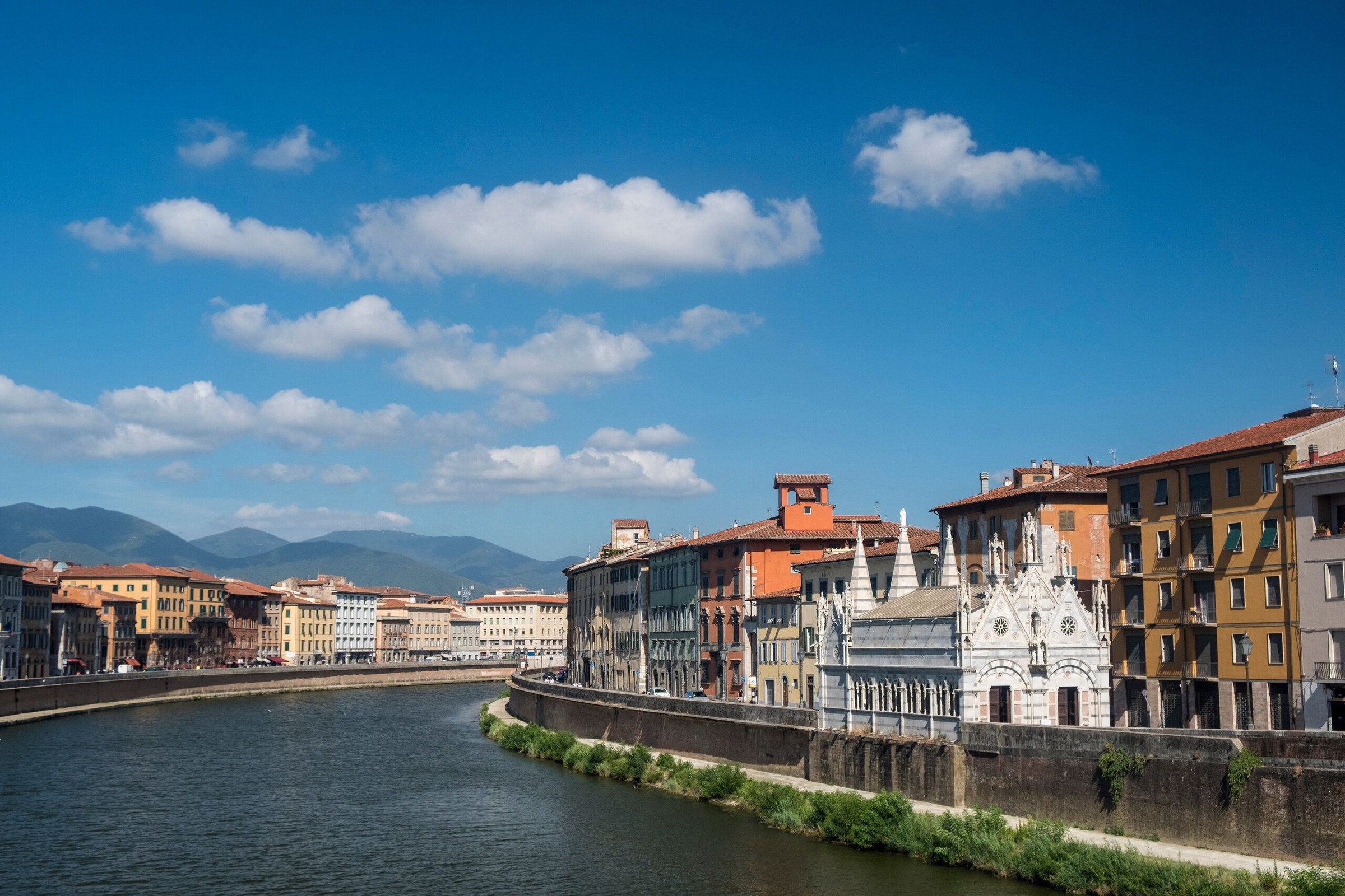 Sungai Arno di Pisa siang hari dengan gereja putih Santa Maria della Spina, deretan rumah warna-warni, dan pegunungan di kejauhan di bawah langit biru berawan.
