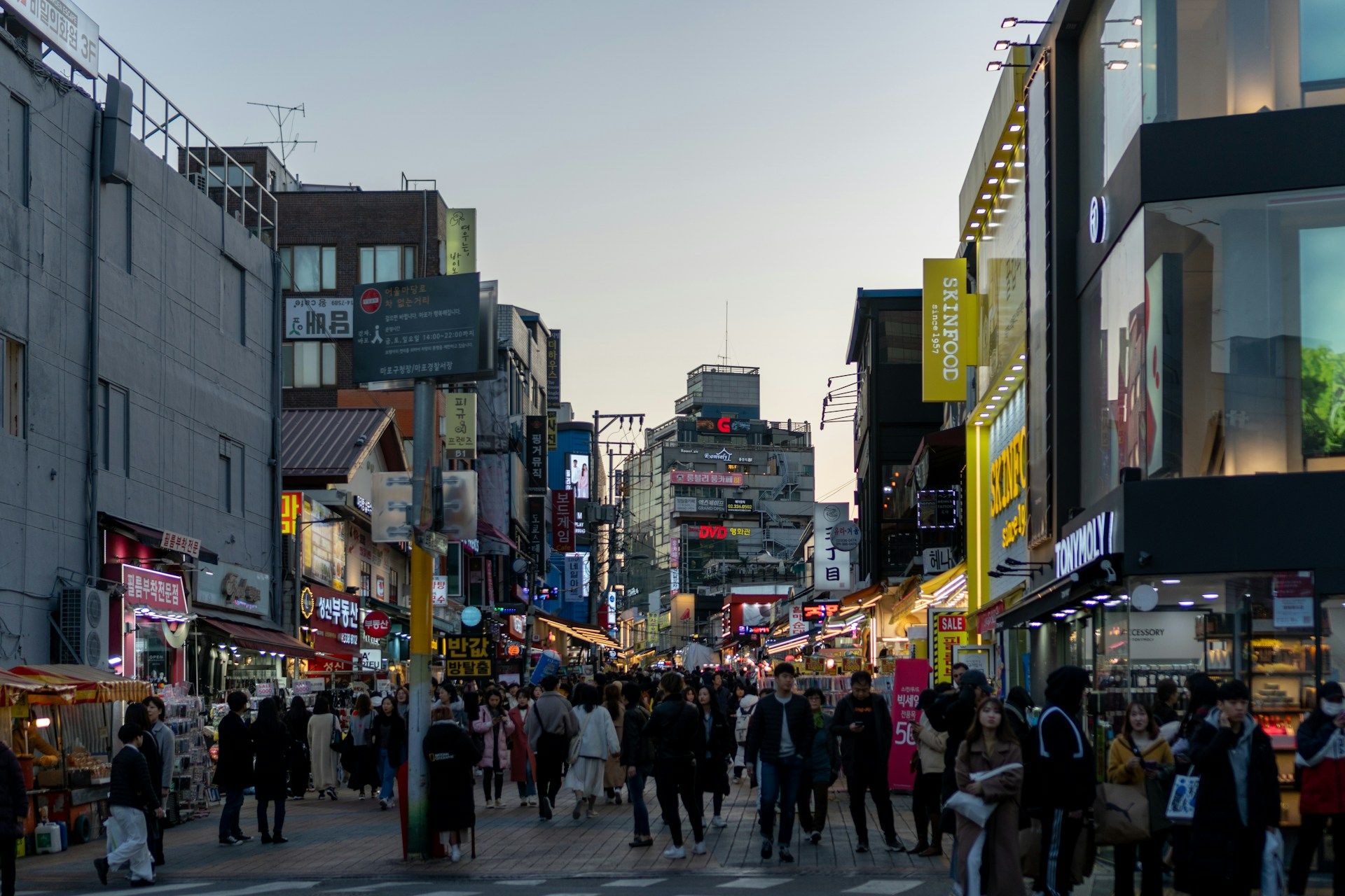 Suasana Hongdae Seoul saat senja, jalan pejalan kaki dipenuhi anak muda, etalase SKINFOOD dan TONYMOLY, signage hangul Korea