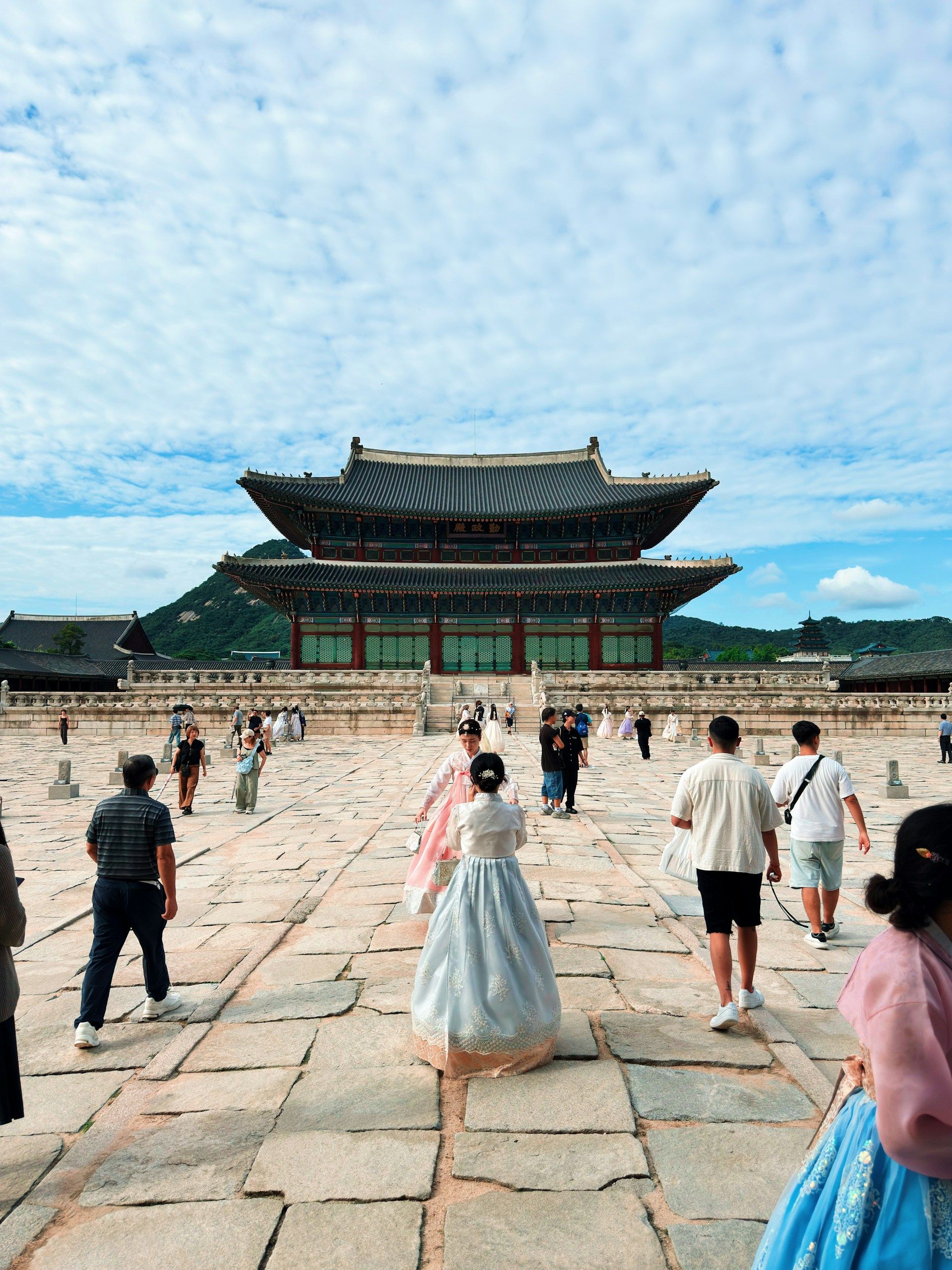 Wisatawan mengenakan hanbok tradisional berwarna pastel berjalan di halaman Istana Gyeongbokgung dengan latar gerbang kayu klasik di musim gugur