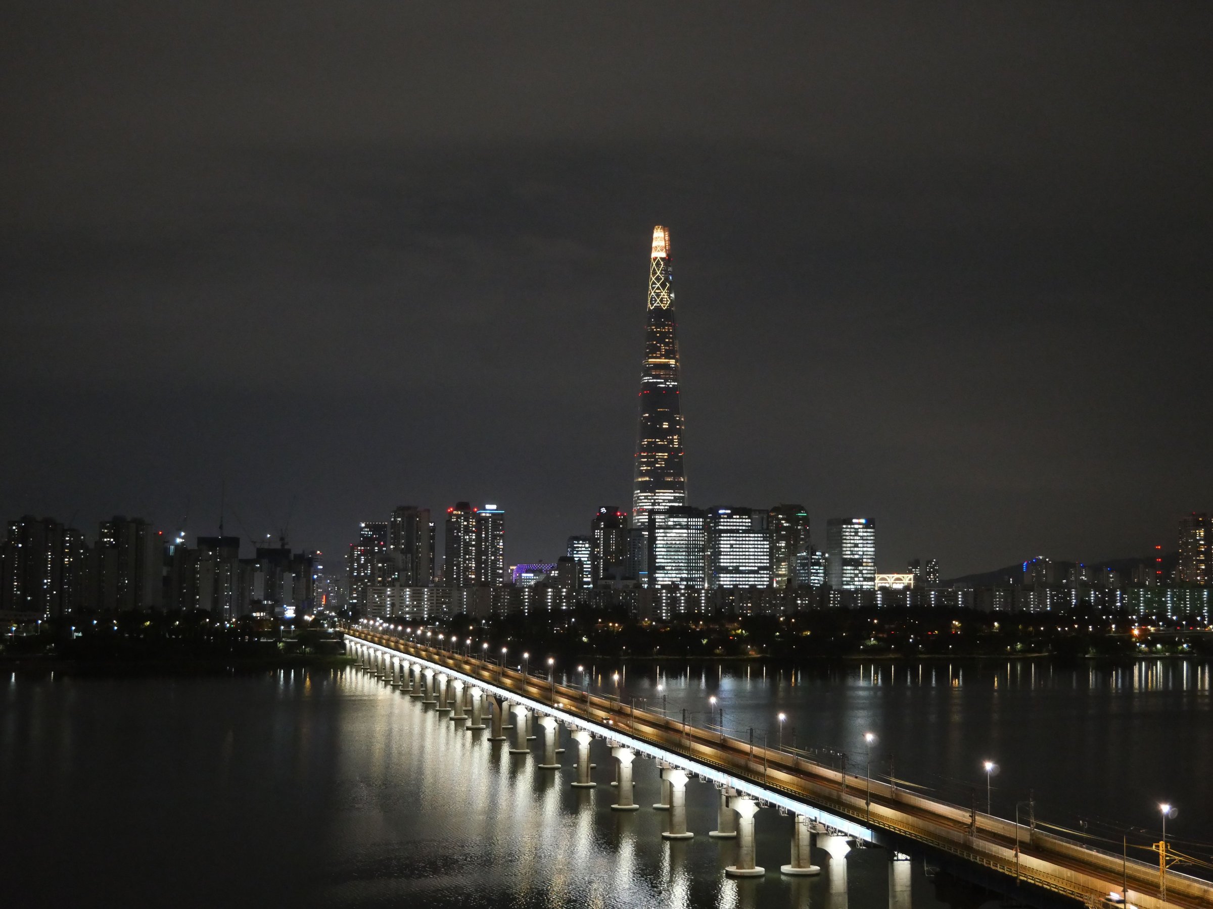 Lotte World Tower menyala malam hari di atas Sungai Han dengan jembatan lurus bercahaya