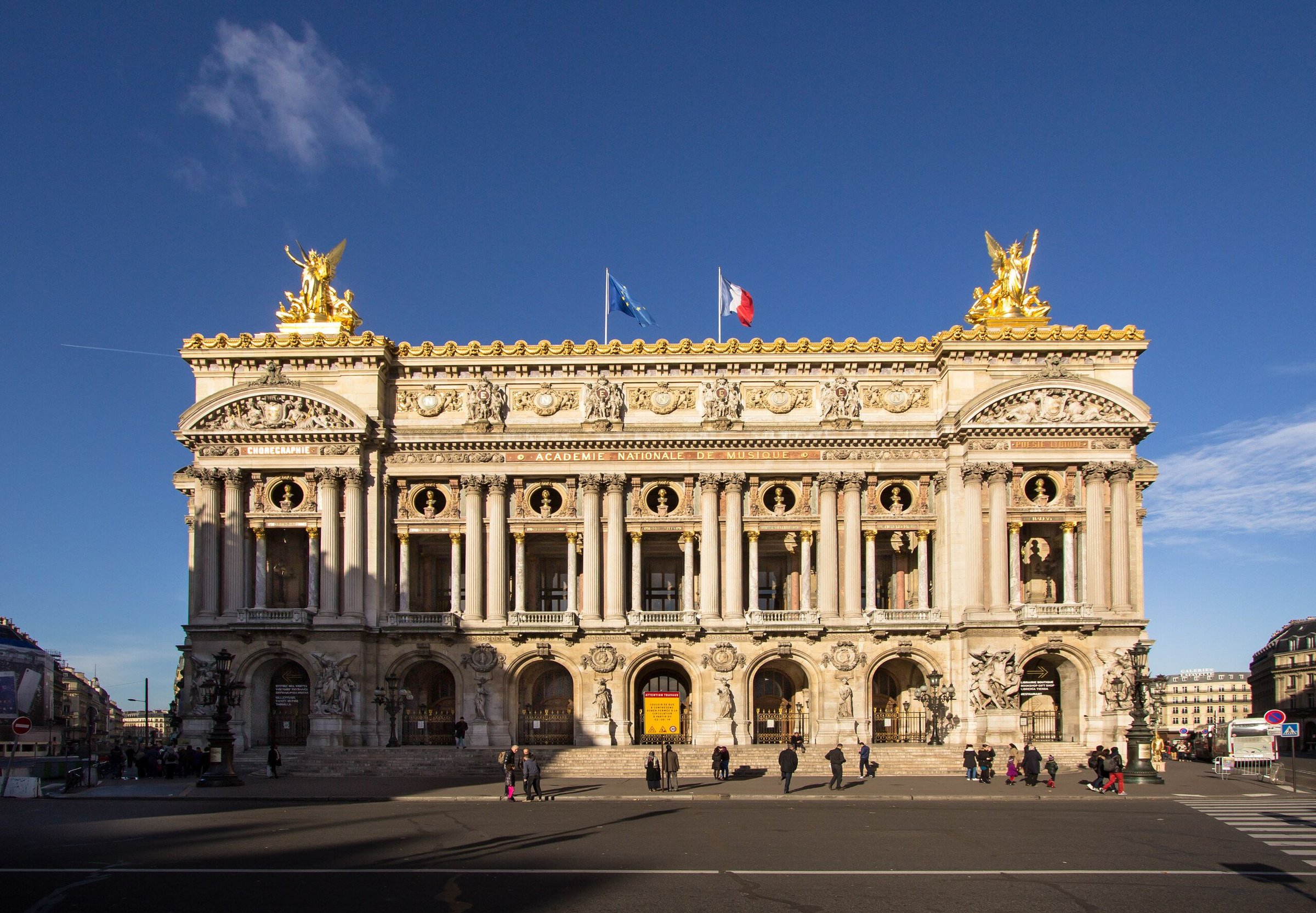 Opéra Garnier di Paris dengan fasad berhias emas, bendera, dan pejalan kaki kecil di plaza saat langit biru cerah.
