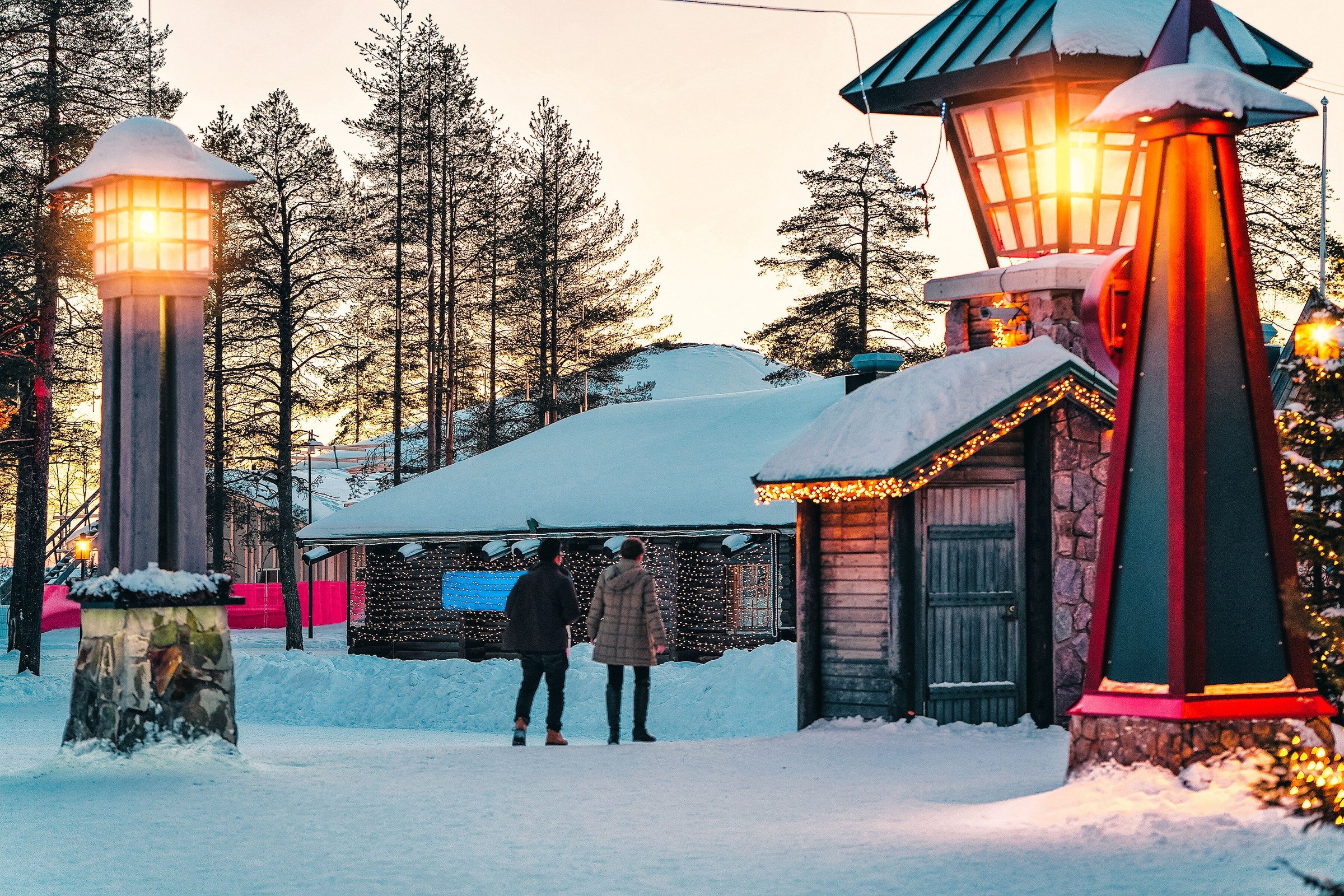 Pasangan berjalan di Santa Claus Village, Rovaniemi, yang bersalju dengan lentera menyala.