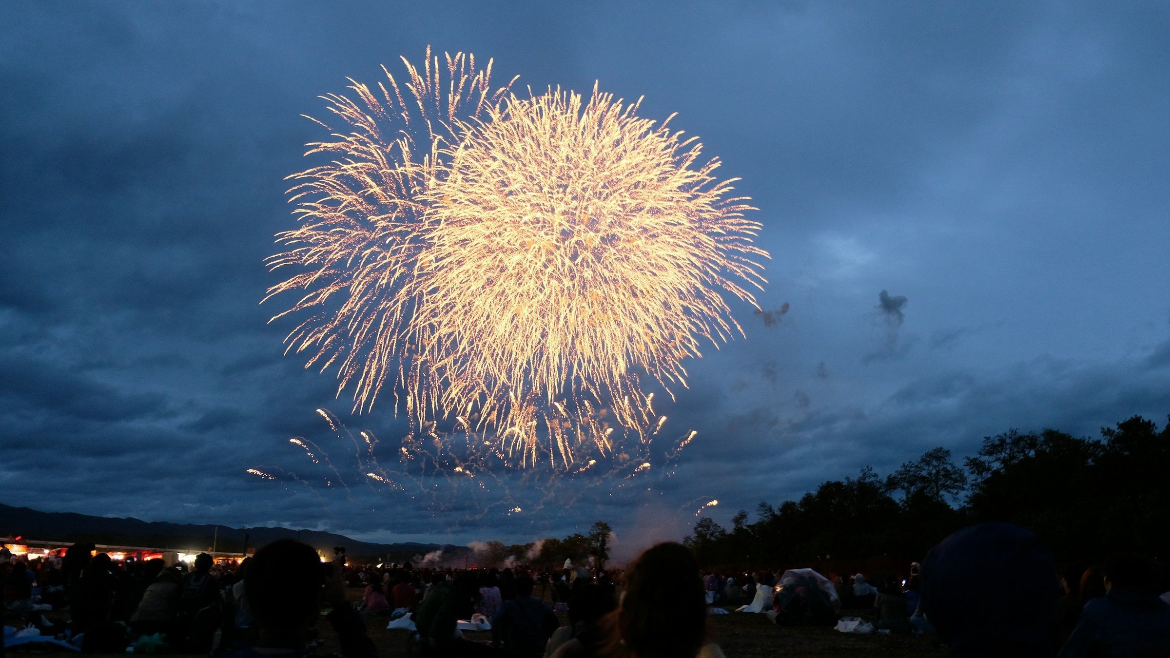 Kembang api hanabi besar berwarna emas merekah di langit senja di atas siluet kerumunan penonton festival musim panas di Hirosaki, Jepang