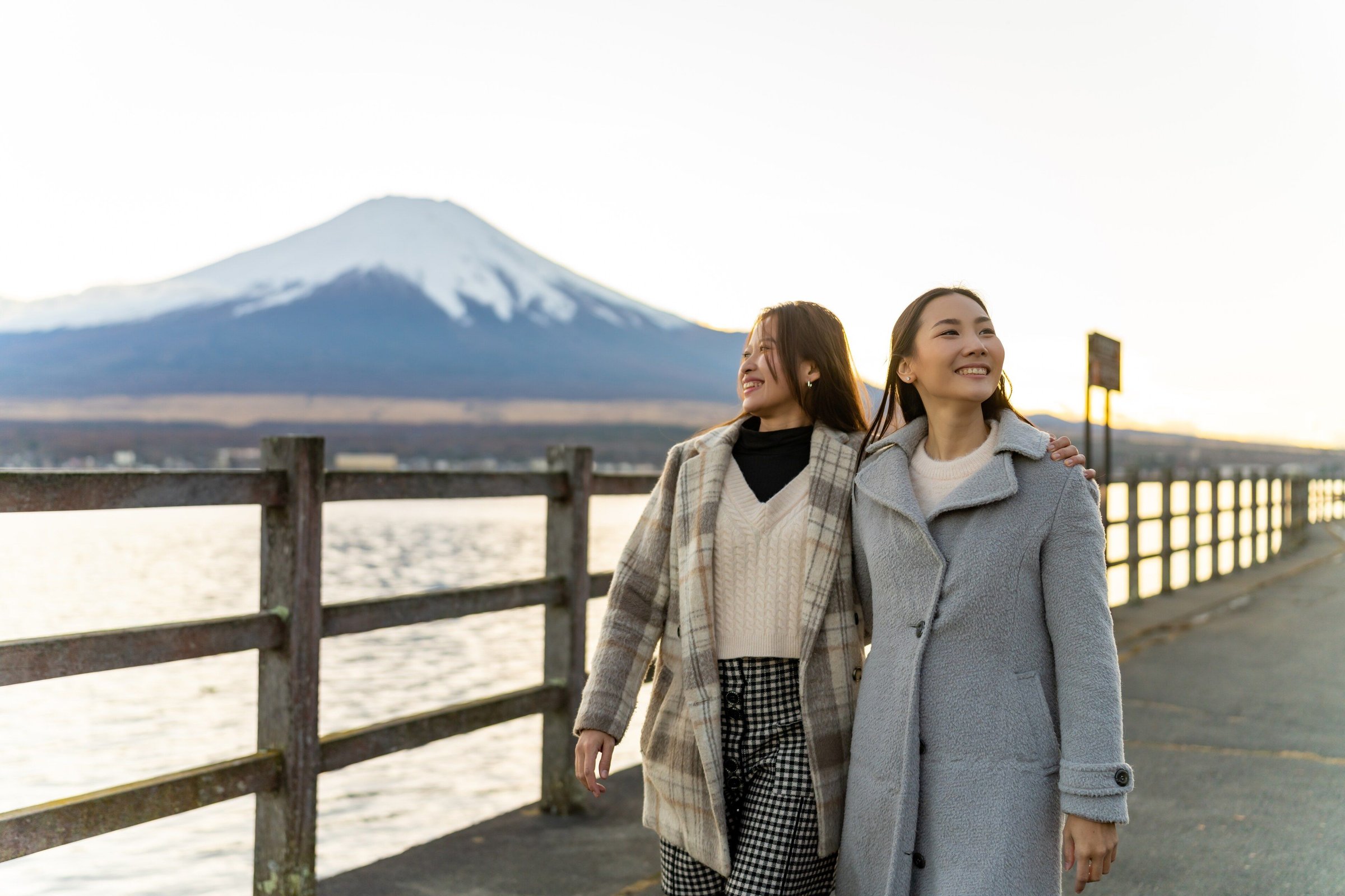 Dua perempuan berjalan di tepi Danau Kawaguchi dengan Gunung Fuji bersalju di latar saat senja