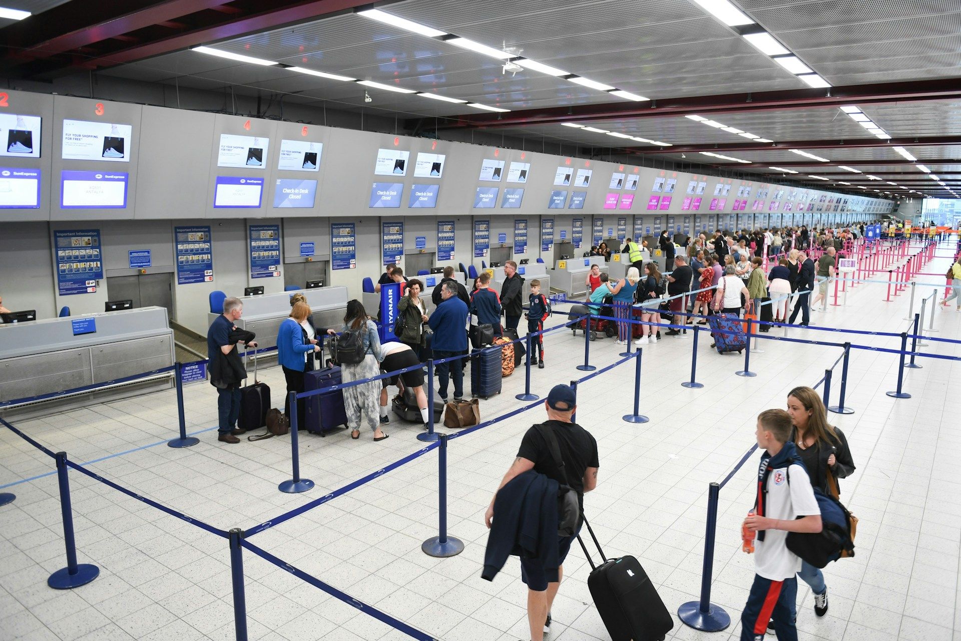 Interior terminal bandara — area check-in dengan deretan counter, antrean stanchion biru, dan traveler membawa koper di hari kepulangan