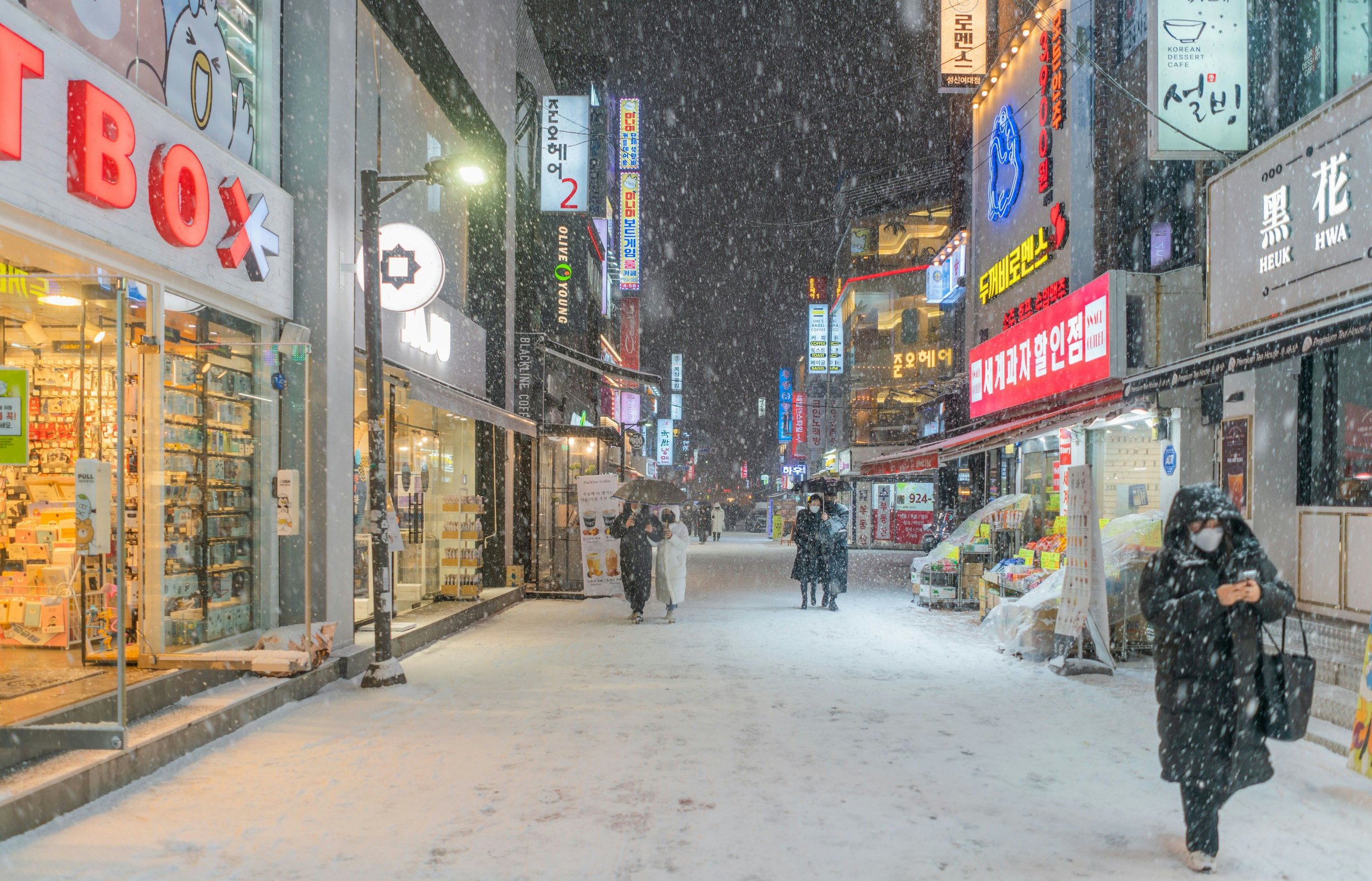 Jalan perbelanjaan Seoul saat salju turun, papan neon menyala dan orang berjalan menembus salju.