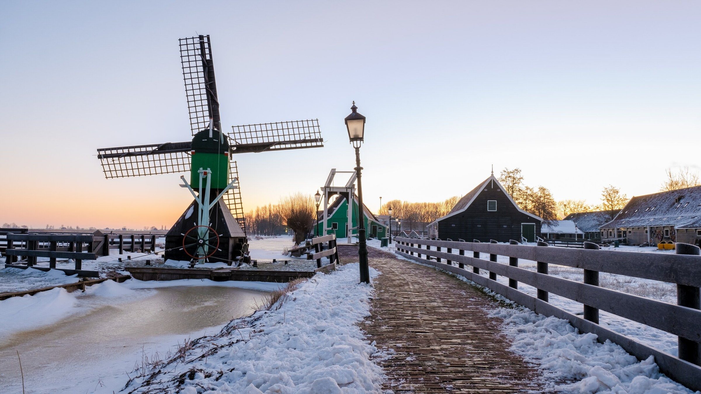 Kincir angin hijau dan lampu jalan di Zaanse Schans dengan salju di jalan setapak dan kanal.