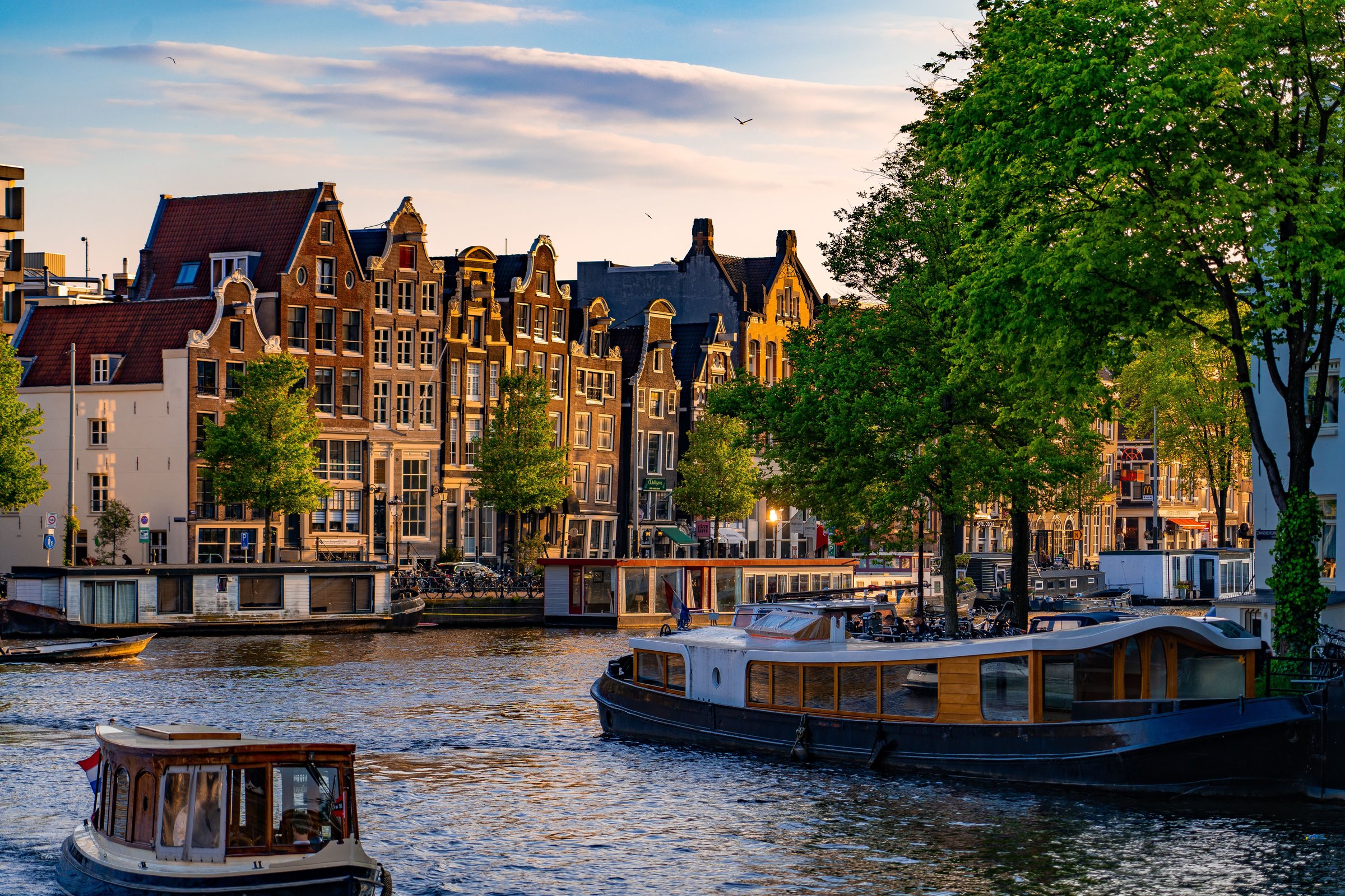 Rumah kanal bergaya gable klasik Amsterdam disinari cahaya keemasan senja dengan kapal wisata