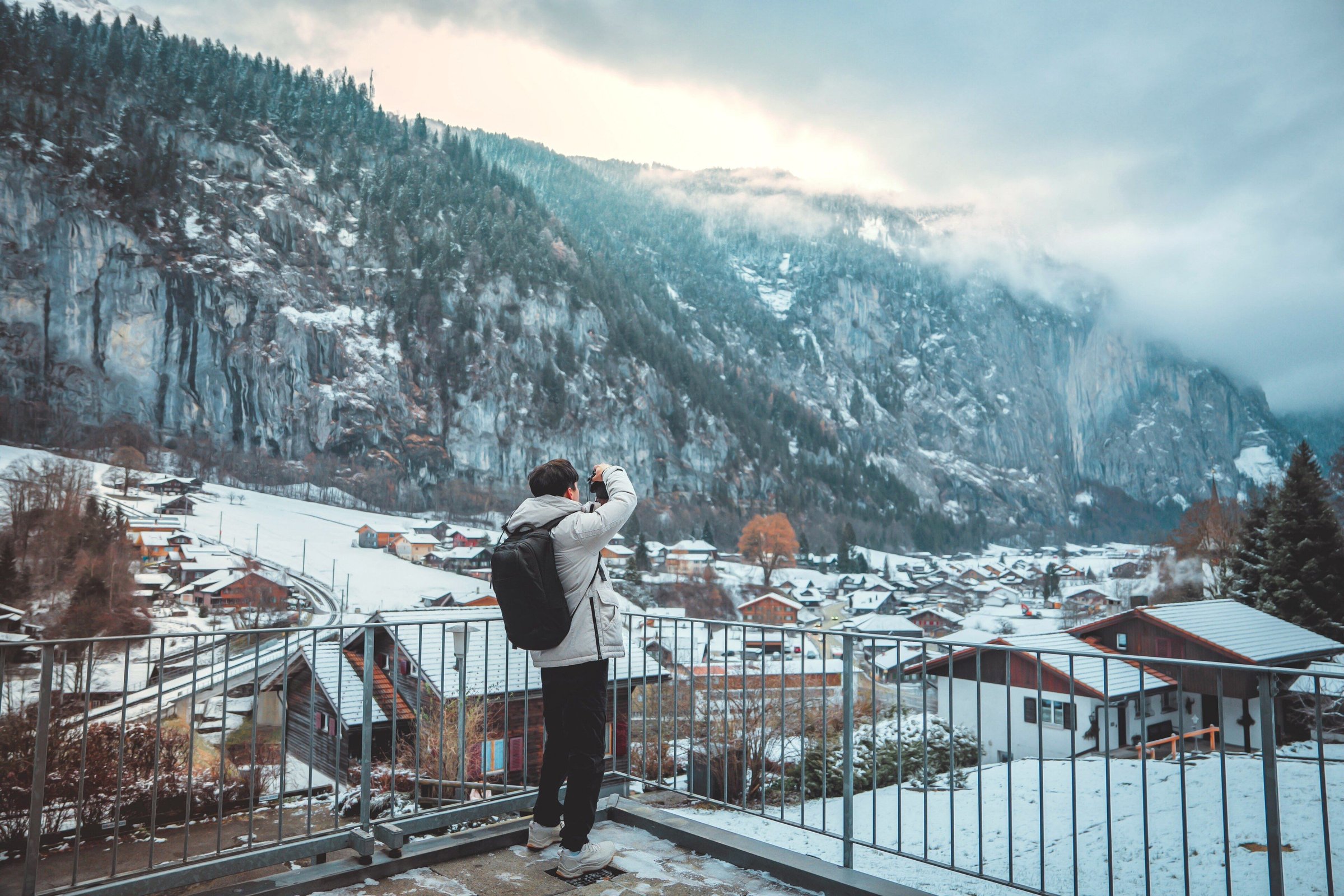Seorang wisatawan berjaket hangat dan membawa ransel memotret desa Lauterbrunnen yang berselimut salju dari pagar teras, tebing berkabut di belakangnya.
