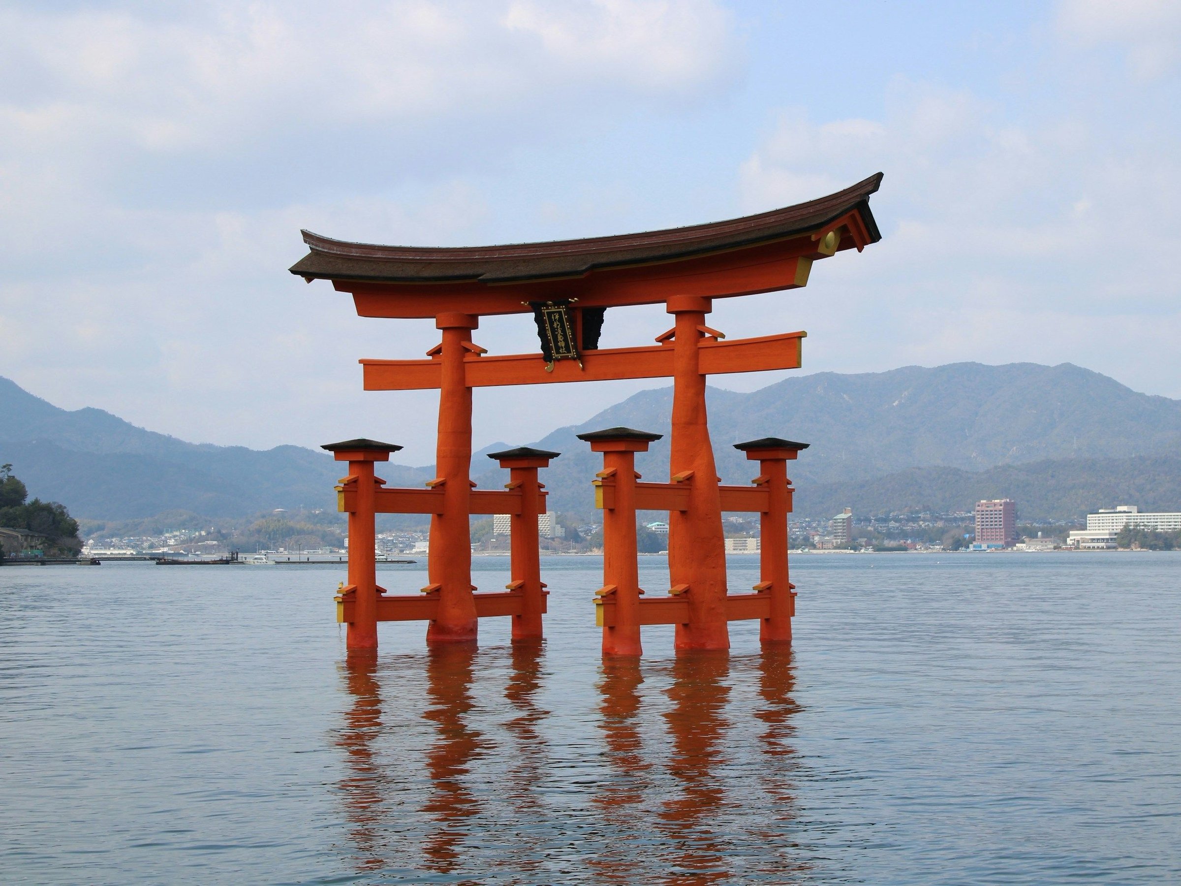 Torii merah Itsukushima mengapung di laut dengan langit cerah dan pegunungan