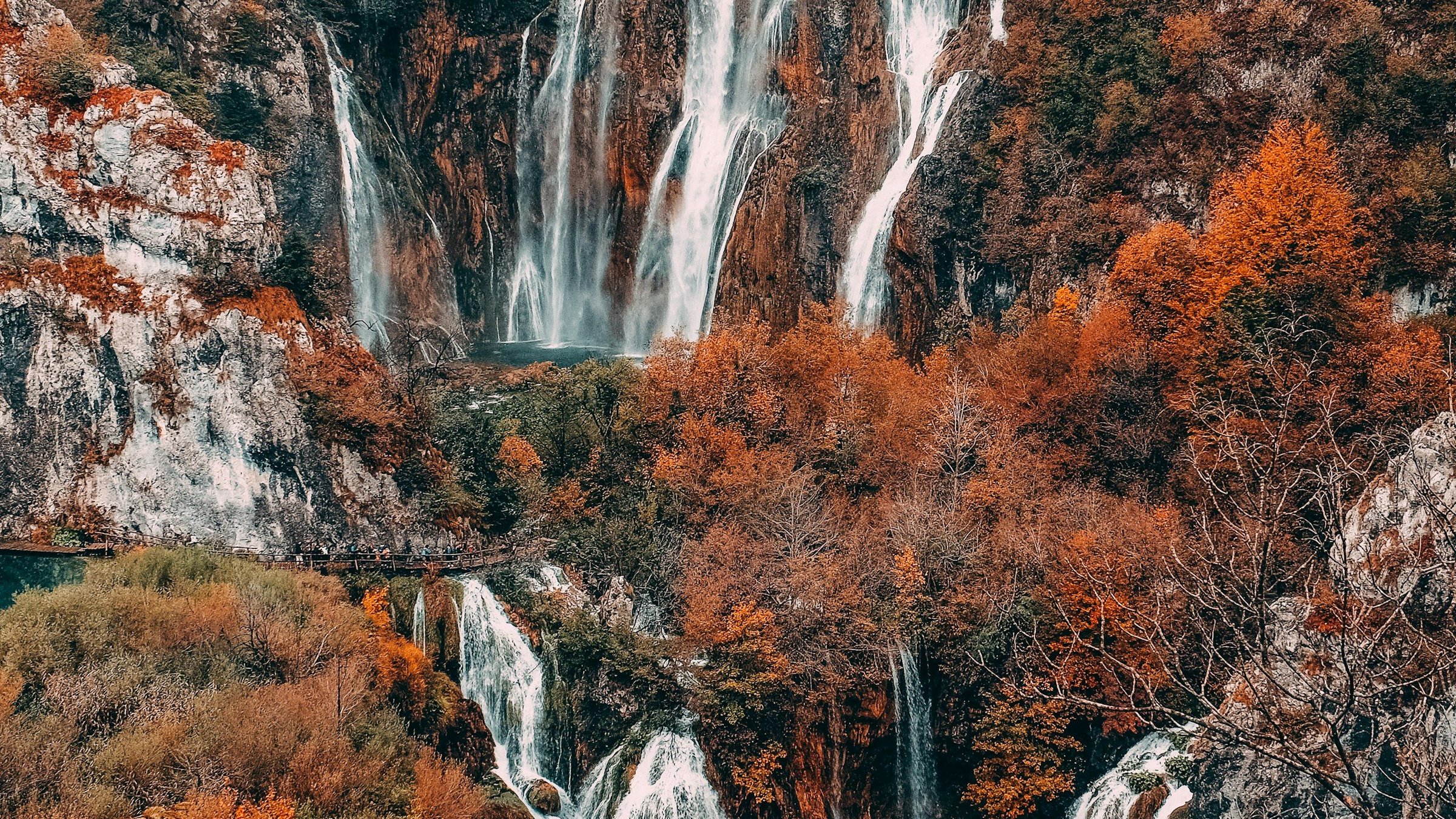 Air terjun Taman Nasional Plitvice di Kroasia dikelilingi dedaunan musim gugur oranye dan merah, dengan kolam berwarna toska dan jalur kayu di bawahnya