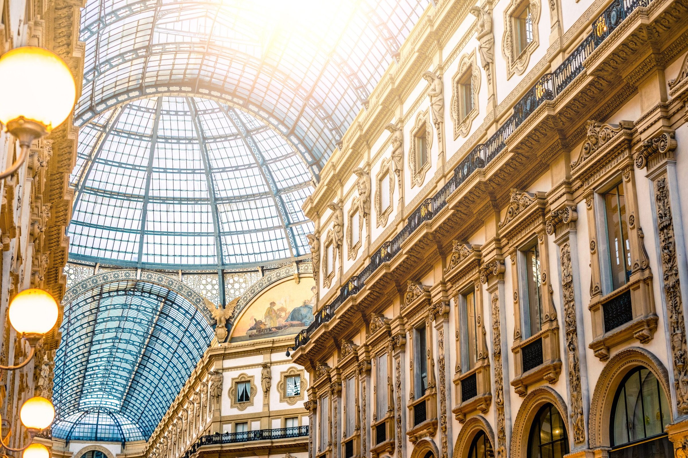 Kubah kaca dan fasad berhias emas di dalam Galleria Vittorio Emanuele II, Milan, dengan cahaya matahari masuk.