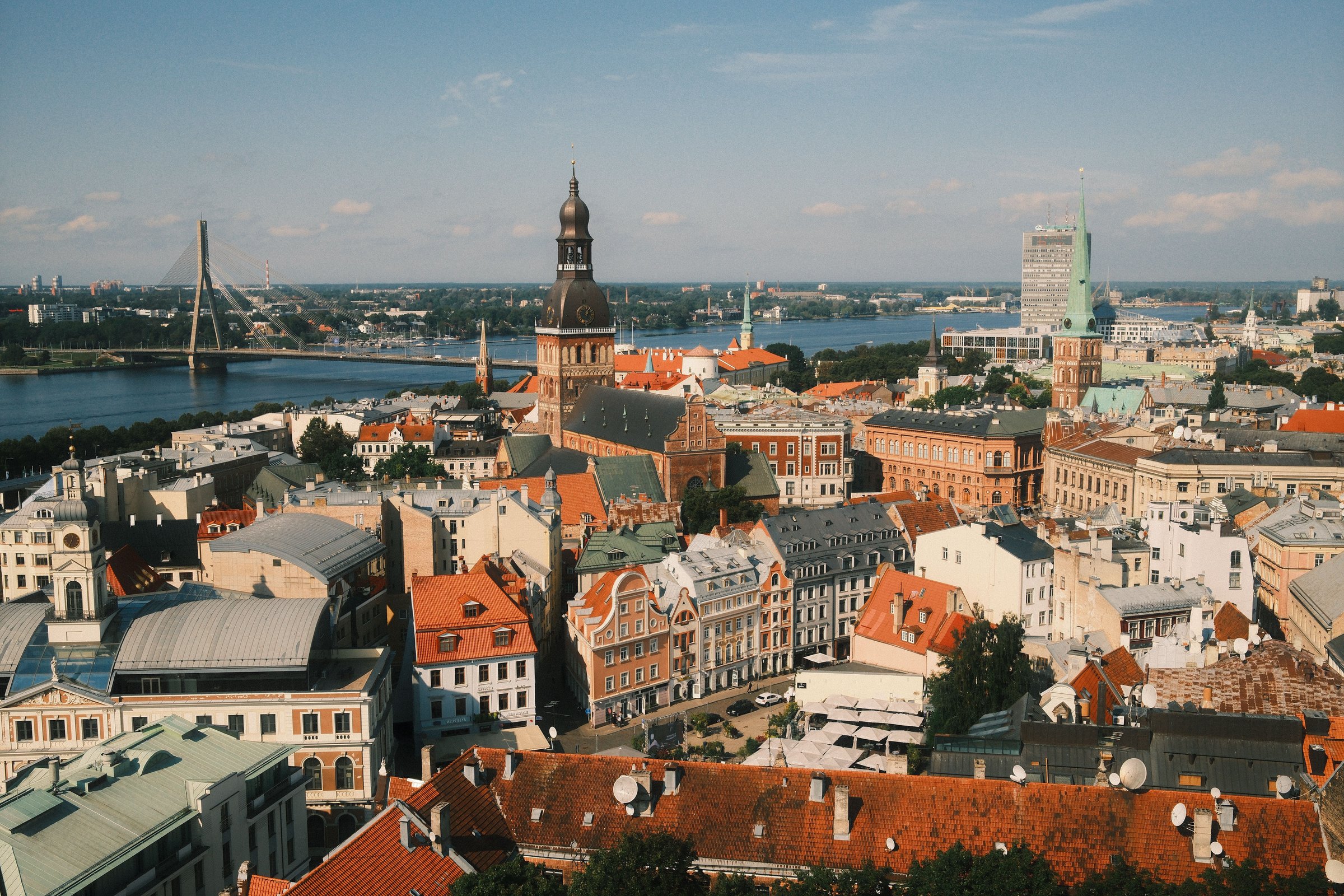 Panorama kota tua Riga dengan menara katedral, sungai, dan atap-atap oranye di bawah langit biru