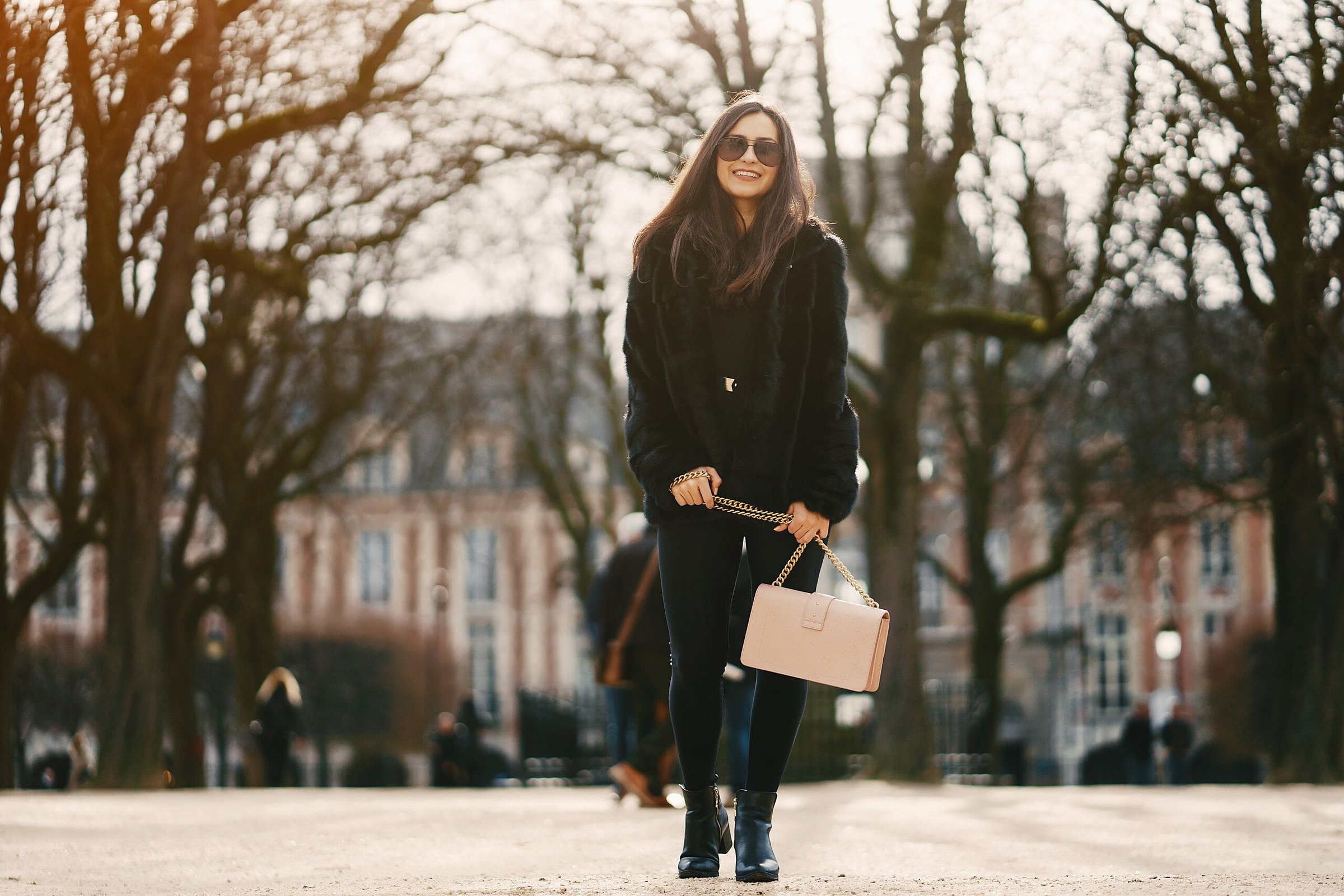 Seorang perempuan berjaket bulu hitam berjalan sambil tersenyum di taman Paris dengan pohon-pohon gundul dan bangunan bata kemerahan di belakangnya.