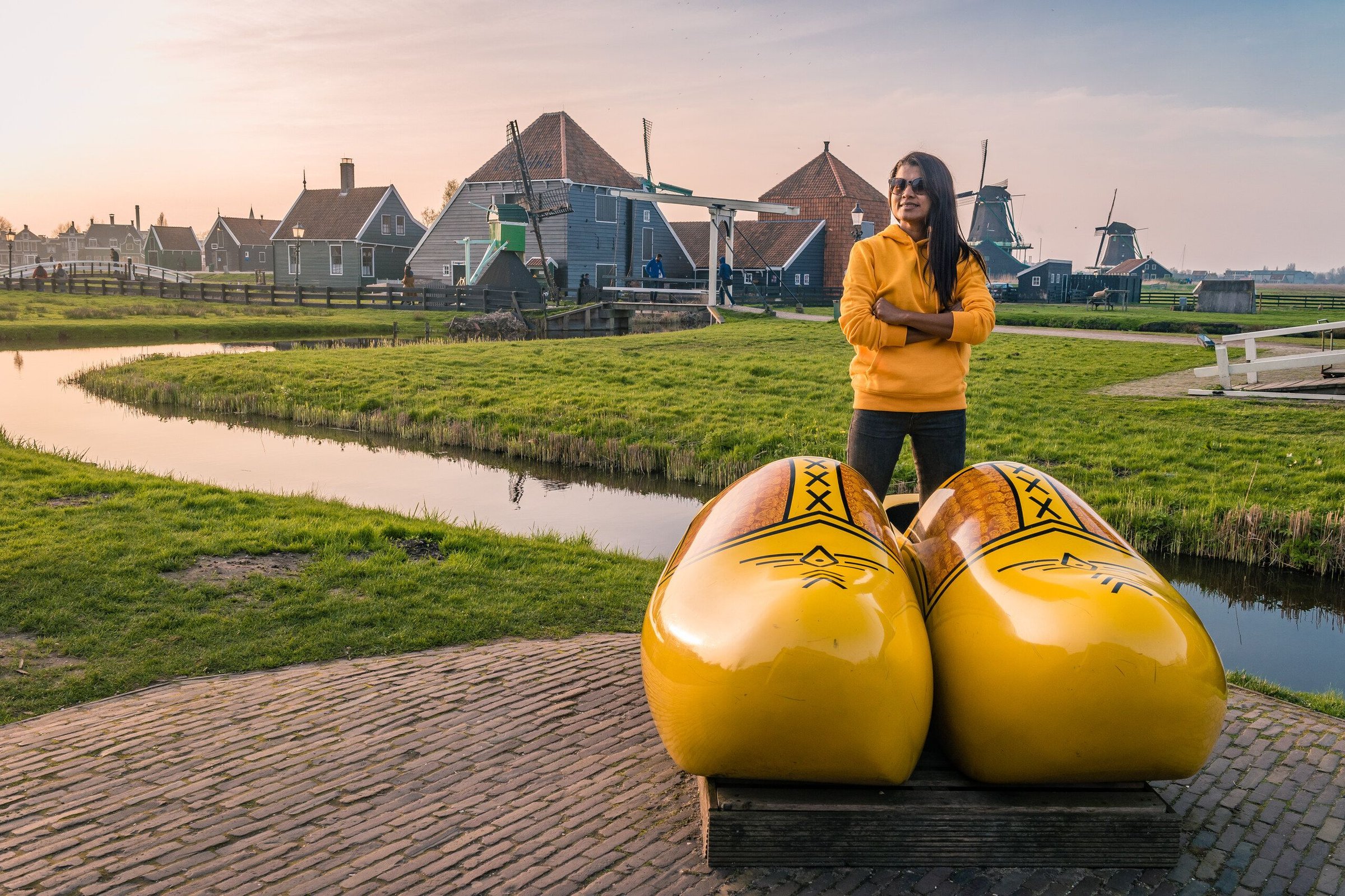 Seorang wisatawan berjaket kuning berdiri di balik sepatu kayu raksasa, dengan kincir angin dan rumah kayu Zaanse Schans di belakangnya.